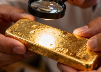 Mains de professionnel examinant une barre d'or avec une loupe.
