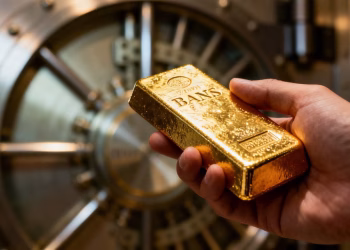 Main d'une personne tenant une barre d'or devant un coffre de banque.
