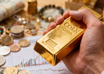 Mano dorada sosteniendo una barra de oro, varios activos financieros en el fondo.